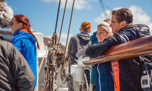 Gemeinsame Momente an Deck Mehrere Menschen stehen an Bord eines Segelschiffs, genießen die Aussicht und das sonnige Wetter, während sie entspannt am Reling lehnen.